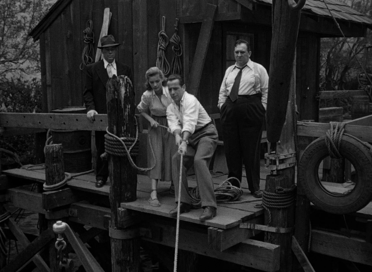 Key Largo (1948) movie still: fullbody — A group of people stands on a weathered wooden pier by a small shed. A man in a white shirt pulls hard on a…