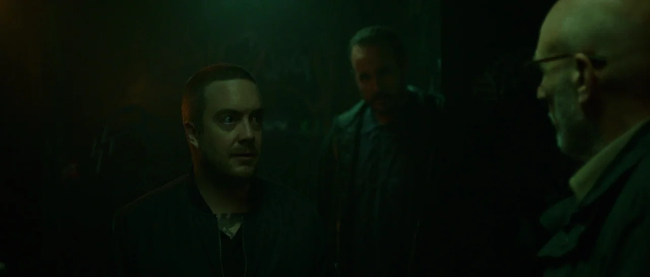 Green Room (2015) movie still: medium — Three men stand in a dark, dimly lit environment with graffiti on the walls. The scene is illuminated by a…