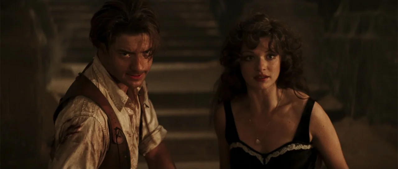 The Mummy (1999) movie still: medium — A stone-walled interior with dark, indistinct architectural elements extending into the shadows behind. A man…