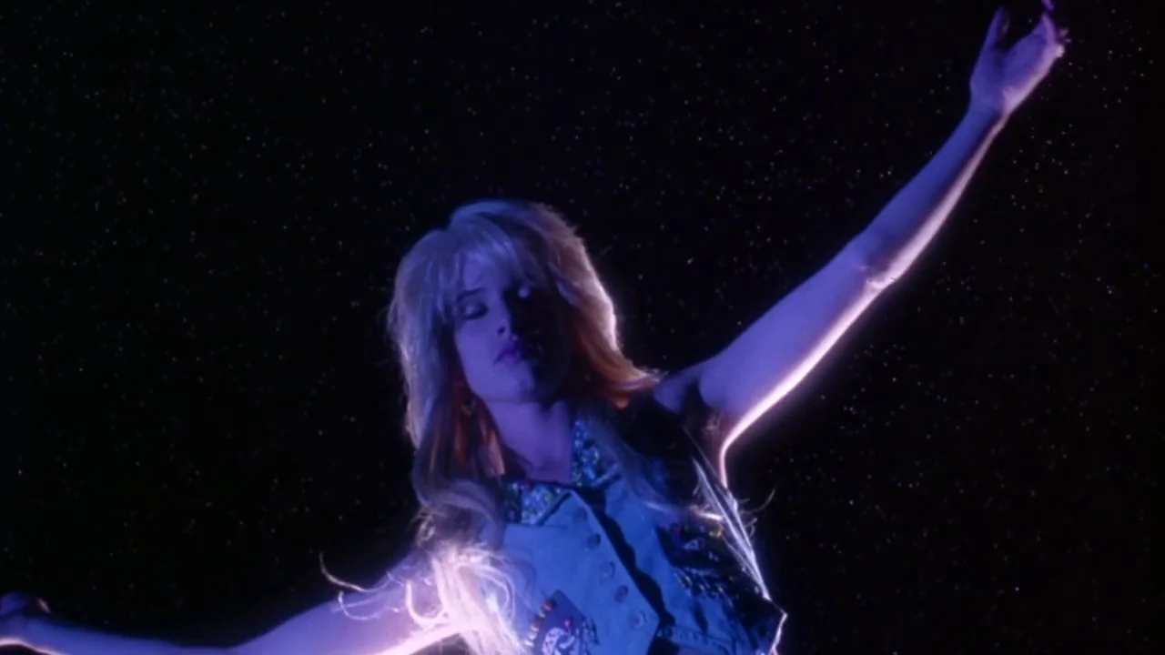 Natural Born Killers (1994) movie still: medium — A young woman stands against a backdrop resembling a dark, starry night sky. She reaches her arms…
