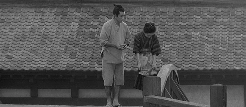 Red Beard (1965) movie still: fullbody — A man and a woman stand on a traditional wooden arched bridge. Behind them, a long building with a tiled…