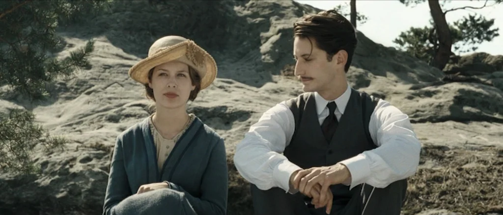Frantz (2016) movie still: medium — A pair of individuals sit together upon a grey rocky outcrop with distant pine trees. The woman in a hat gazes…