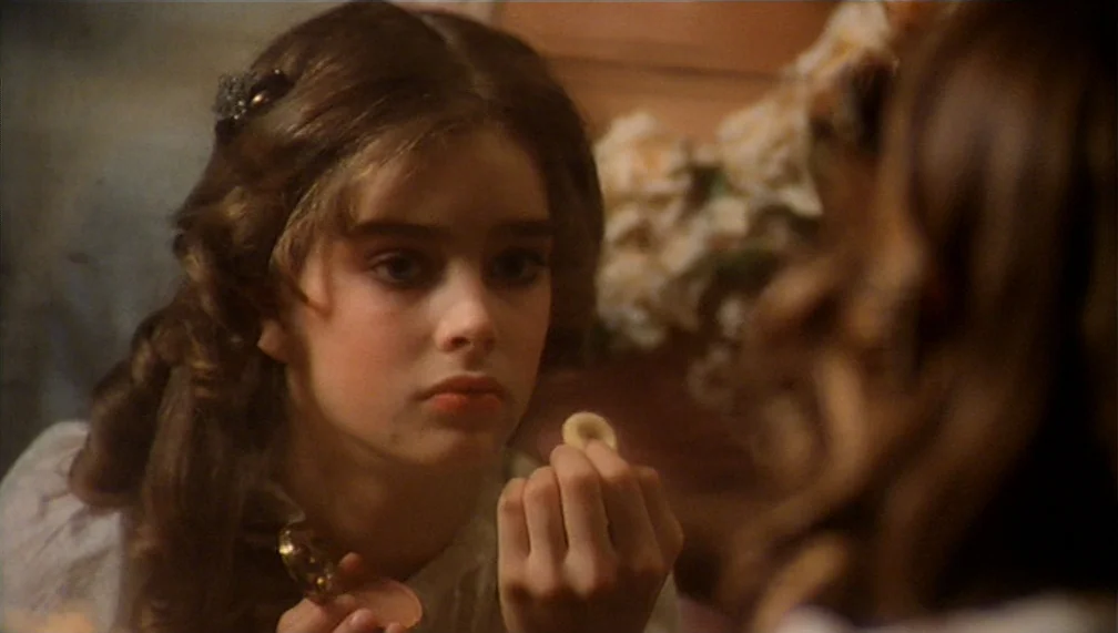 Pretty Baby (1978) movie still: closeup — A young girl with wavy brown hair looks into a reflective glass surface. She holds a small round powder…