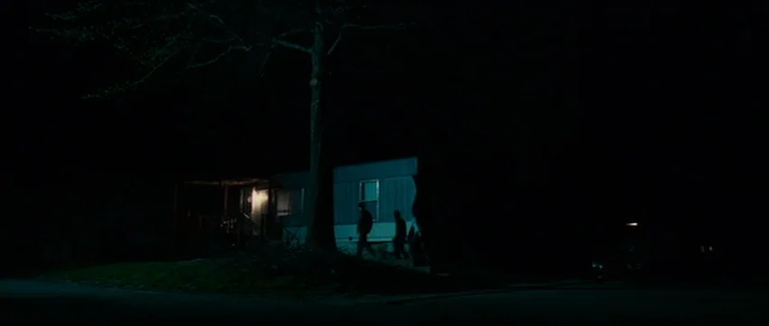 Bubble (2005) movie still: wide — A dimly lit exterior view of a mobile home at night with a large tree. Three figures walk across the lawn toward…