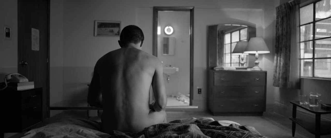Roma (2018) movie still: medium — A sparsely furnished motel room with a patterned bedspread and a wooden dresser. A man sits shirtless on the bed…