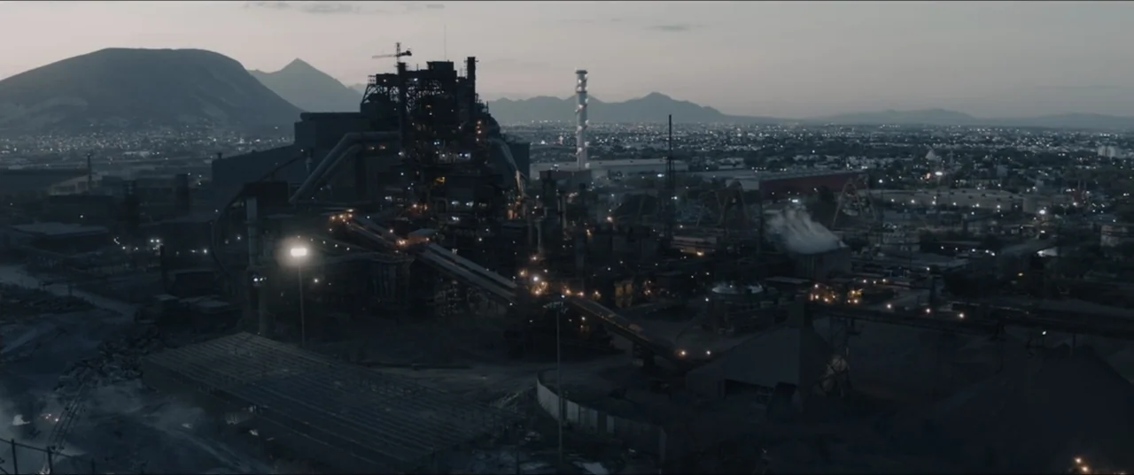 Death to Summer (2021) movie still: establishing — A sprawling industrial steel mill complex stands against a backdrop of mountains and city lights.…