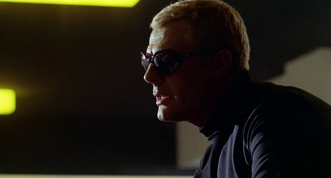 The 10th Victim (1965) movie still: closeup — A man stands in a dark room with a bright yellow light strip behind him. The man is wearing dark…