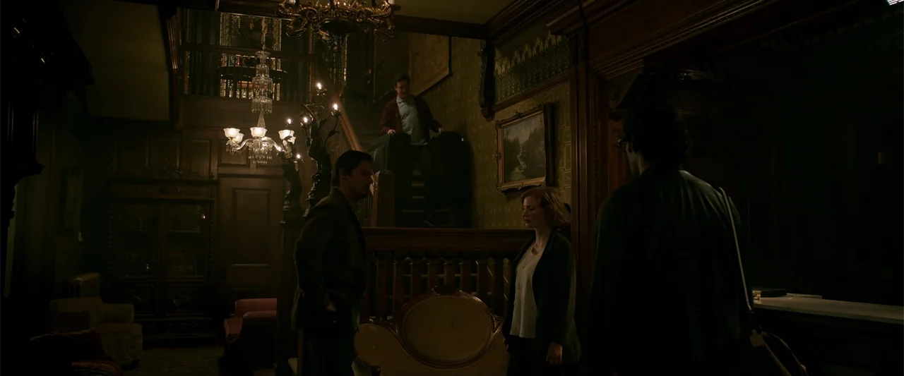 K.G.F: Chapter 2 (2022) movie still: wide — A grand, dark Victorian mansion foyer with a wooden staircase and crystal chandeliers. Three individuals…