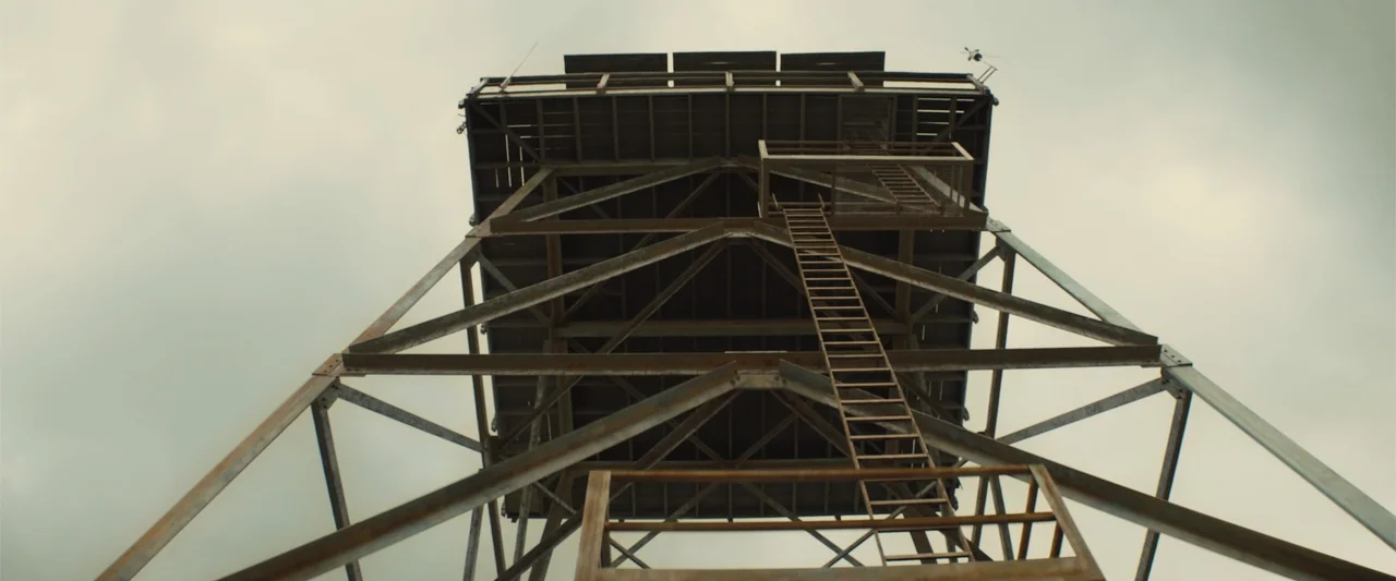 Those Who Wish Me Dead (2021) movie still: establishing — A metal fire lookout tower with a ladder stands against an overcast sky. The structure is…
