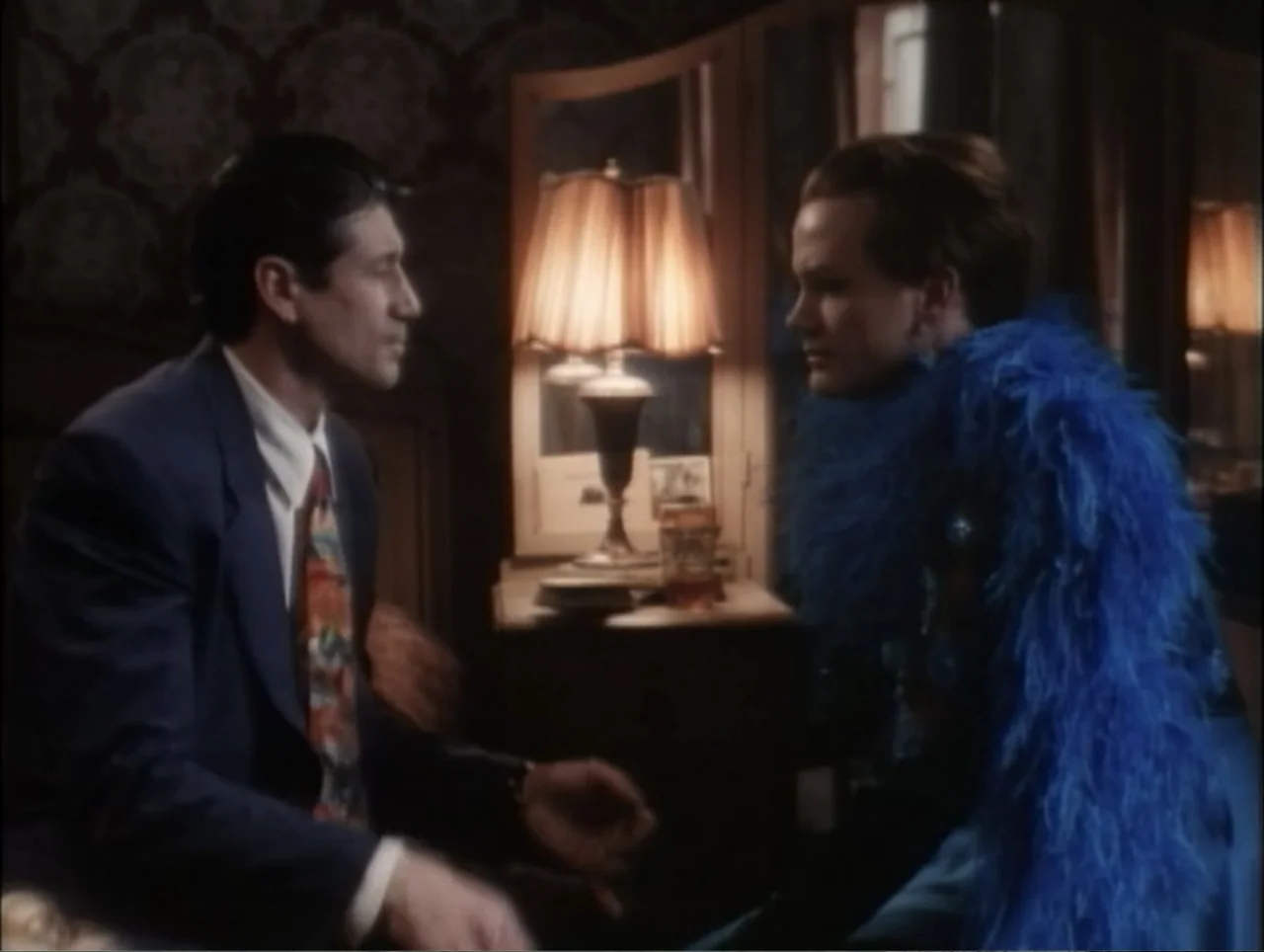 Cast a Deadly Spell (1991) movie still: medium — A dimly lit room with patterned wallpaper features a lit table lamp. A man in a suit sits opposite…