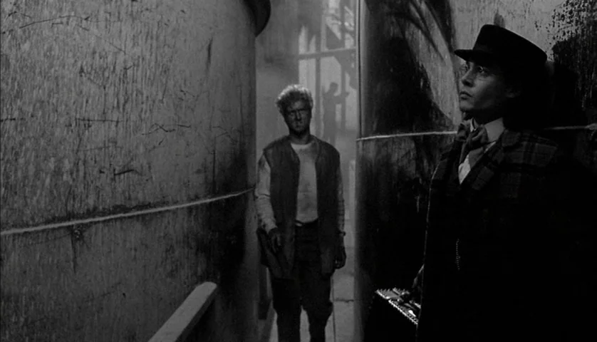 Dead Man (1995) movie still: medium — A narrow corridor flanked by two large, weathered metal storage tanks in a dark facility. A man in a suit…