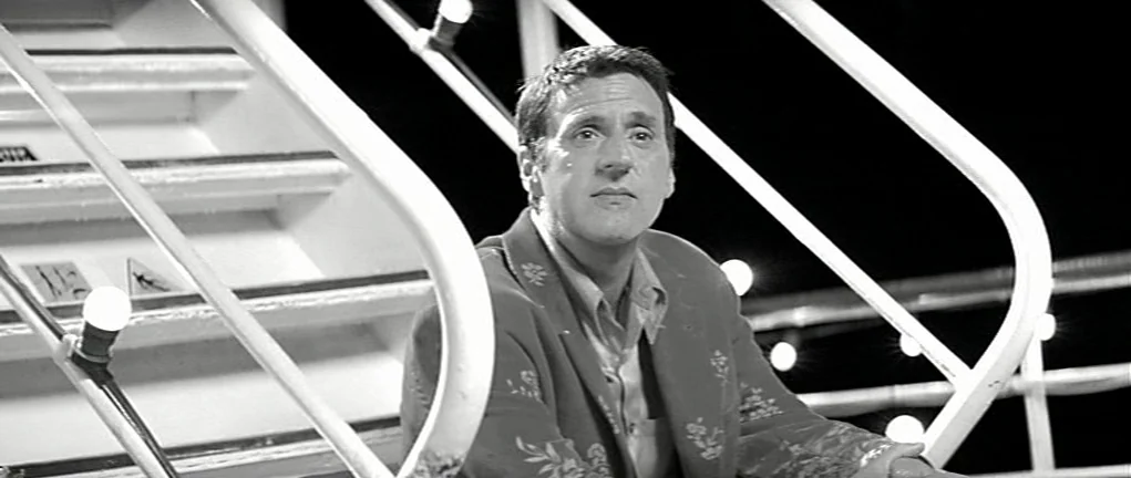 Girl on the Bridge (1999) movie still: medium — A man sits on a metal staircase on the deck of a ship at night. Multiple small white light bulbs are…
