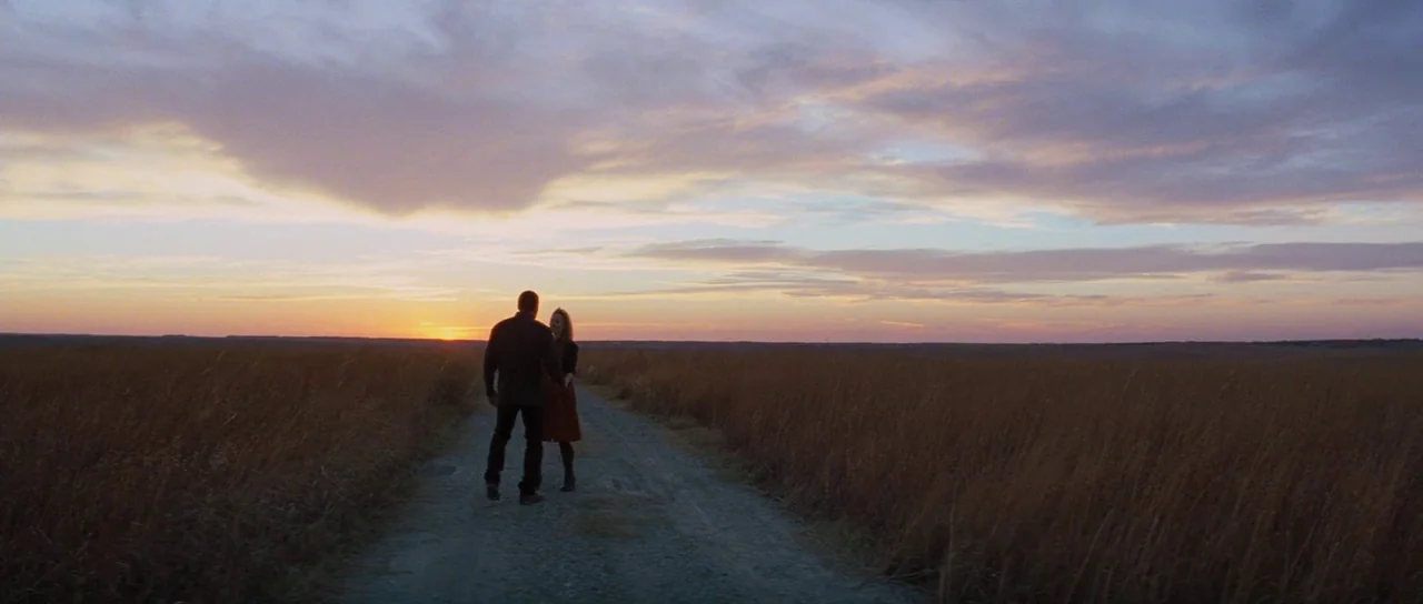 To The Wonder (2012) movie still: wide — A flat prairie landscape under a large sky at golden hour. A man and woman stand on a narrow dirt track in…