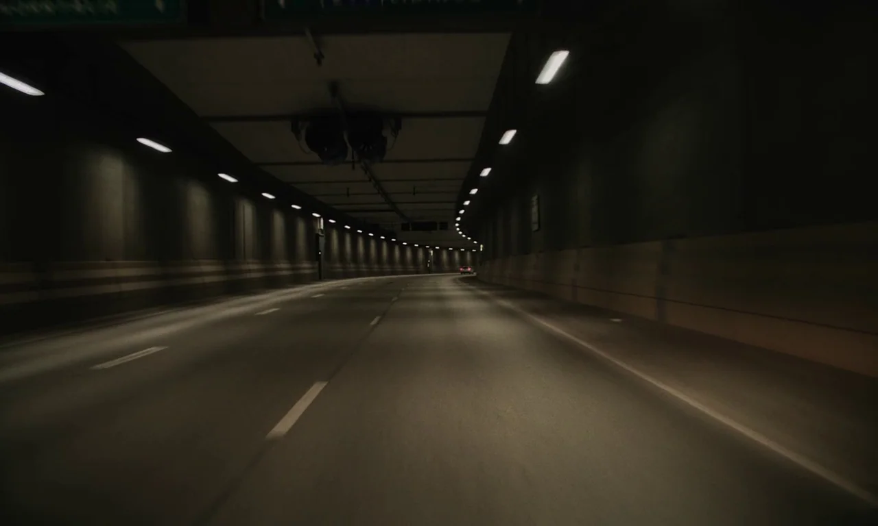 Vox Lux (2018) movie still: establishing — A long, curved concrete road tunnel stretches forward into the dark, vanishing point. A single vehicle…