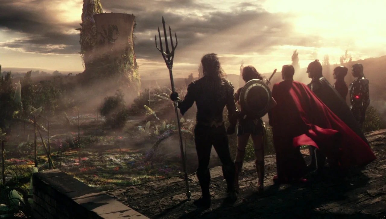 Justice League (2017) movie still: wide — A group of six figures stands on a dark rocky edge overlooking a valley. A large ruined tower overgrown…