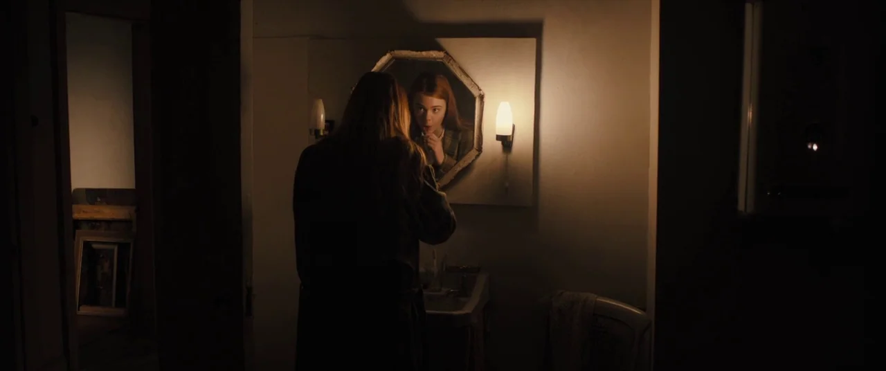 Ginger & Rosa (2012) movie still: medium — A dimly lit room with a wall-mounted mirror flanked by two illuminated light fixtures. A woman with long…