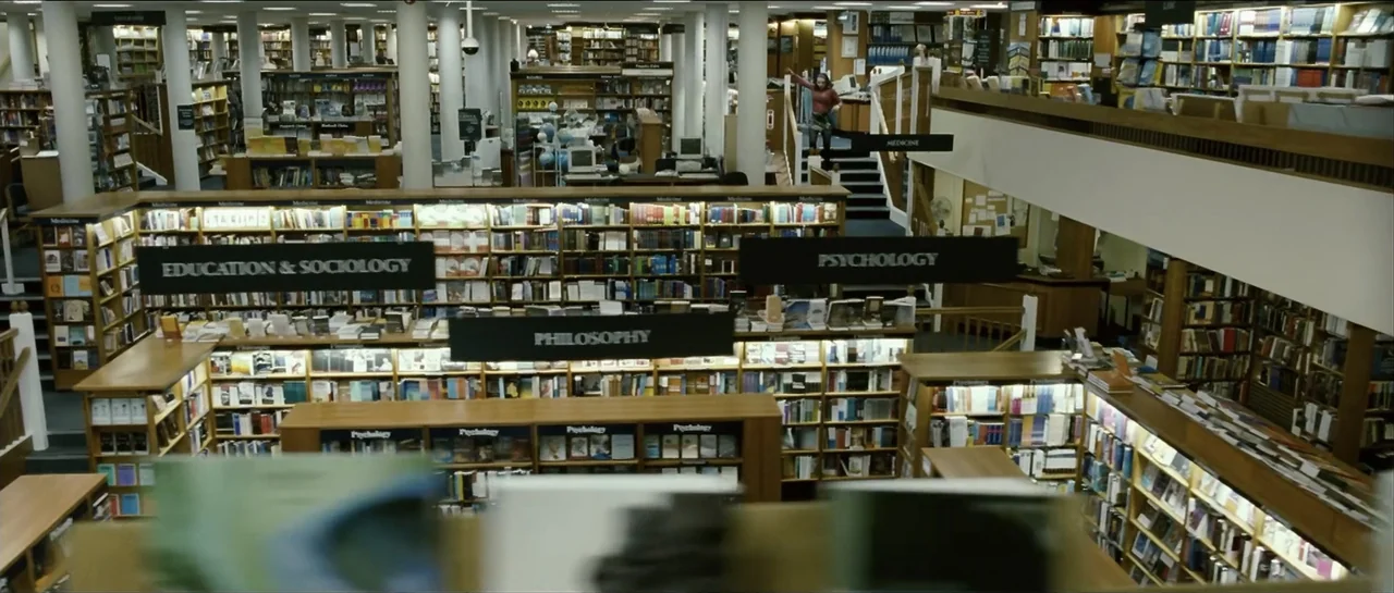 The Oxford Murders (2008) movie still: wide — A multi-level bookstore features expansive wooden bookshelves categorized by genre signage. A person…