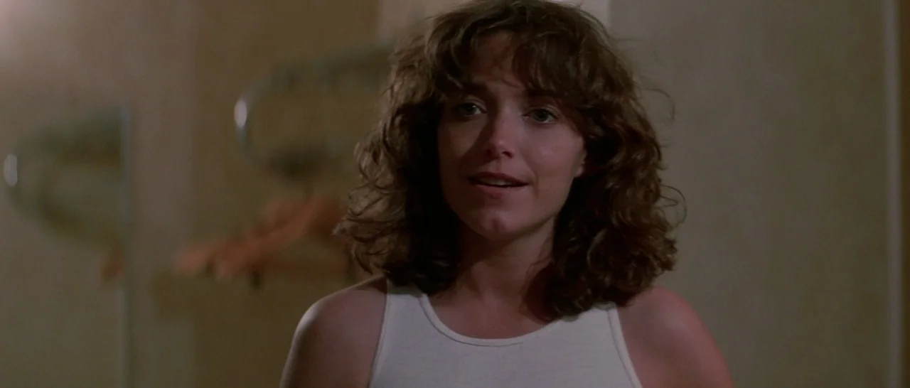 Starman (1984) movie still: medium — A woman stands in a small, neutral-toned space with a wall-mounted garment hook. The woman in a white tank top…