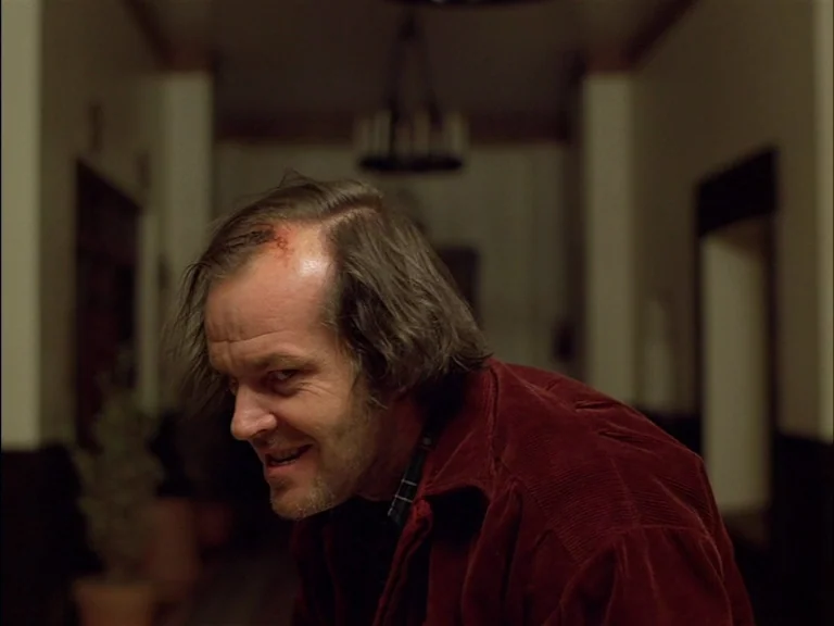 The Shining (1980) movie still: medium — A dimly lit interior hotel corridor features dark wood accents and architectural details. A middle-aged man…