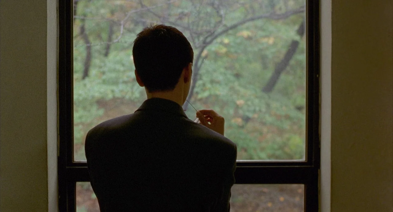 Barking Dogs Never Bite (2000) movie still: medium — A person stands before a large window looking out at a densely wooded area. They hold glasses…
