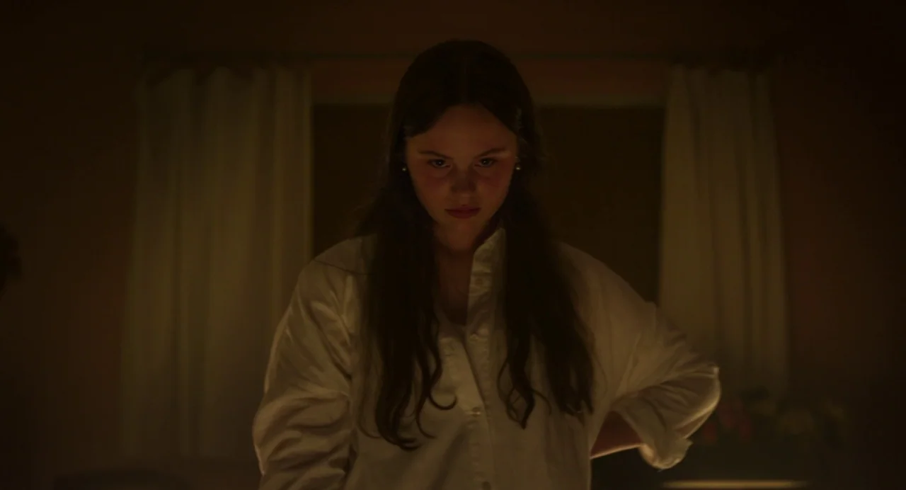 Zombi Child (2019) movie still: medium — A dimly lit room with white curtains visible behind the subject. A woman stands in front of the window…