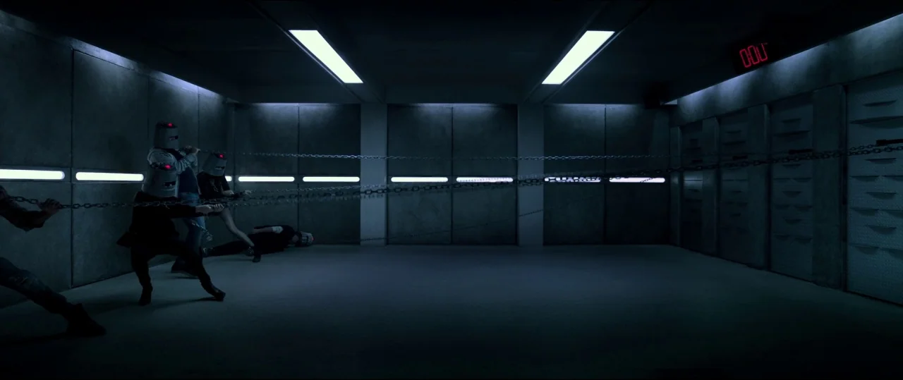 Jigsaw (2017) movie still: wide — An industrial concrete chamber features metallic wall panels and long fluorescent light strips. Several…
