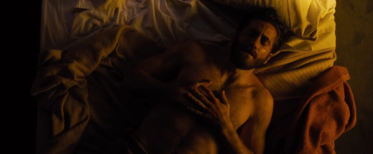 Nocturnal Animals (2016) movie still: medium — A man lies on rumpled white bed sheets under dim amber lighting. He rests with eyes closed while his…