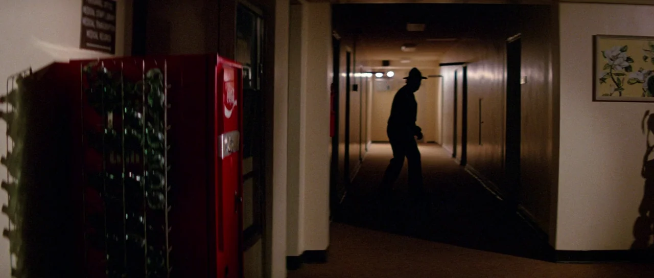 Halloween II (1981) movie still: wide — A long, dimly lit hospital hallway with beige walls and closed doors on both sides. A man in a hat walks…