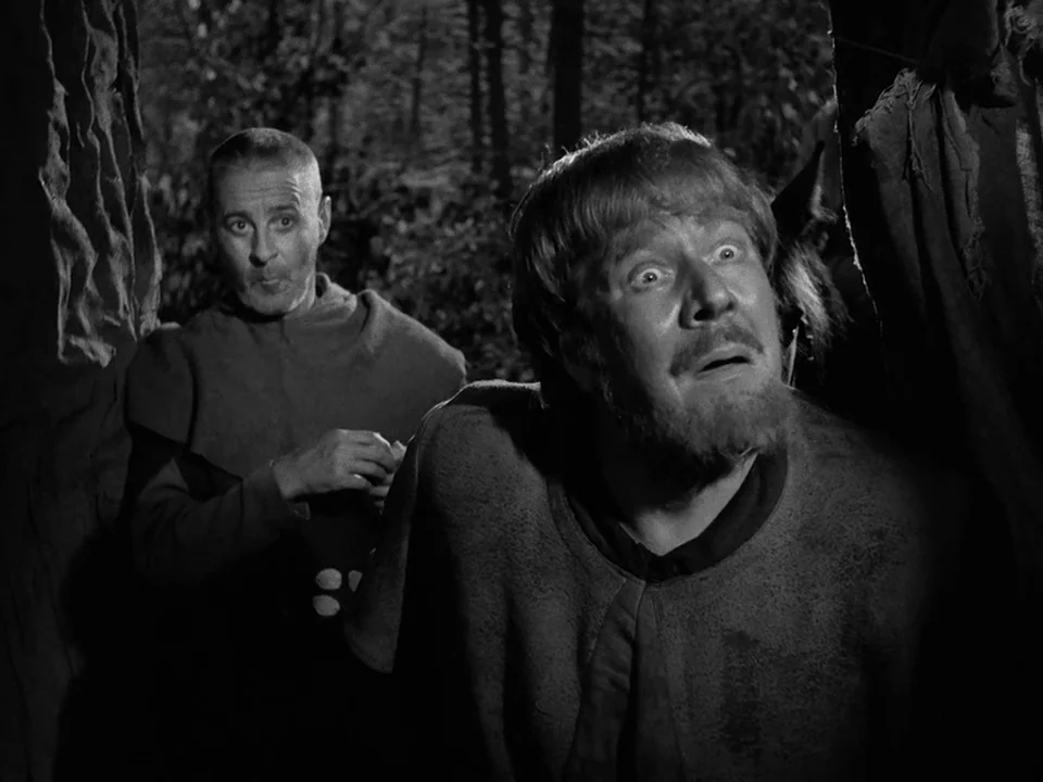 The Seventh Seal (1957) movie still: medium — A dense forest setting with thick tree trunks framing the two foreground subjects. Two men peer…