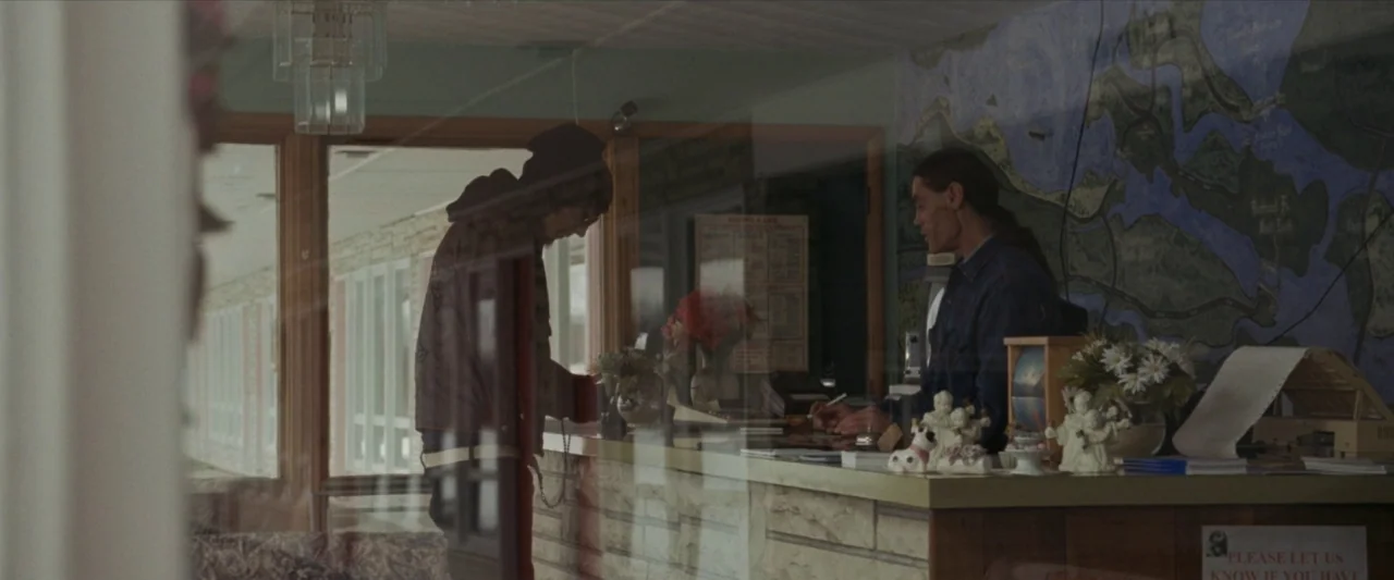 For Ellen (2012) movie still: wide — A motel lobby interior features a wood-paneled counter and a large wall-mounted map. A customer in a hooded…