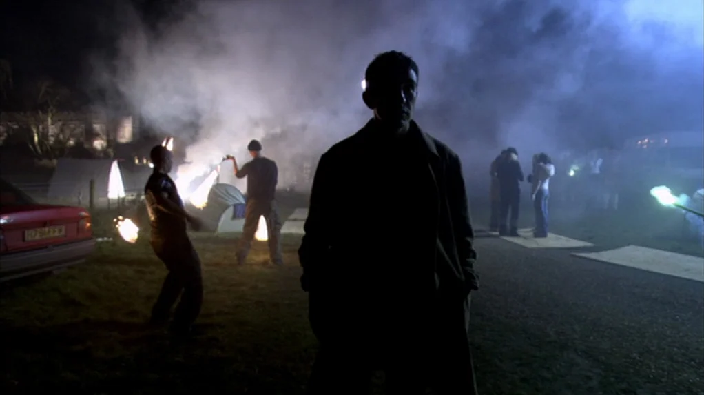 A Cock and Bull Story (2005) movie still: medium — A nighttime outdoor campsite features several people gathered around a small flickering fire.…