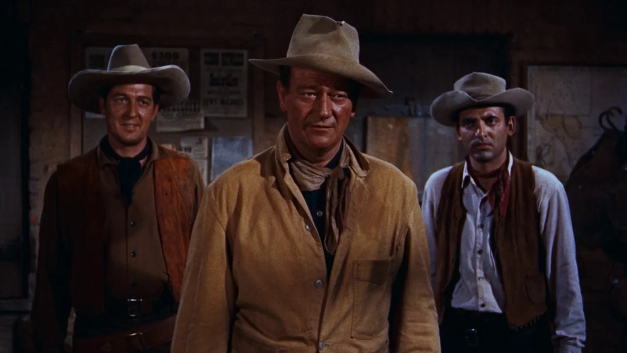 Rio Bravo (1959) movie still: medium — Three men wearing cowboy hats stand in a dimly lit, rustic wooden building interior. They face toward the…
