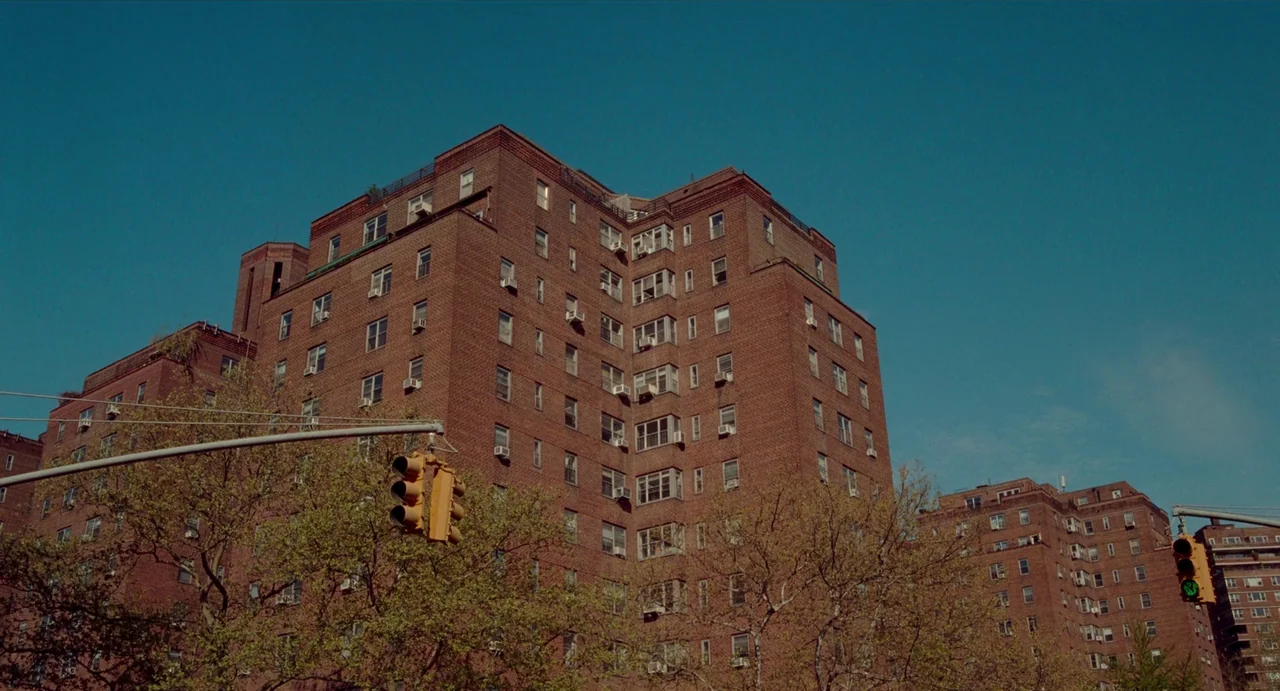 His Three Daughters (2023) movie still: establishing — Tall red brick apartment buildings rise against a clear blue sky. Traffic lights hang on…