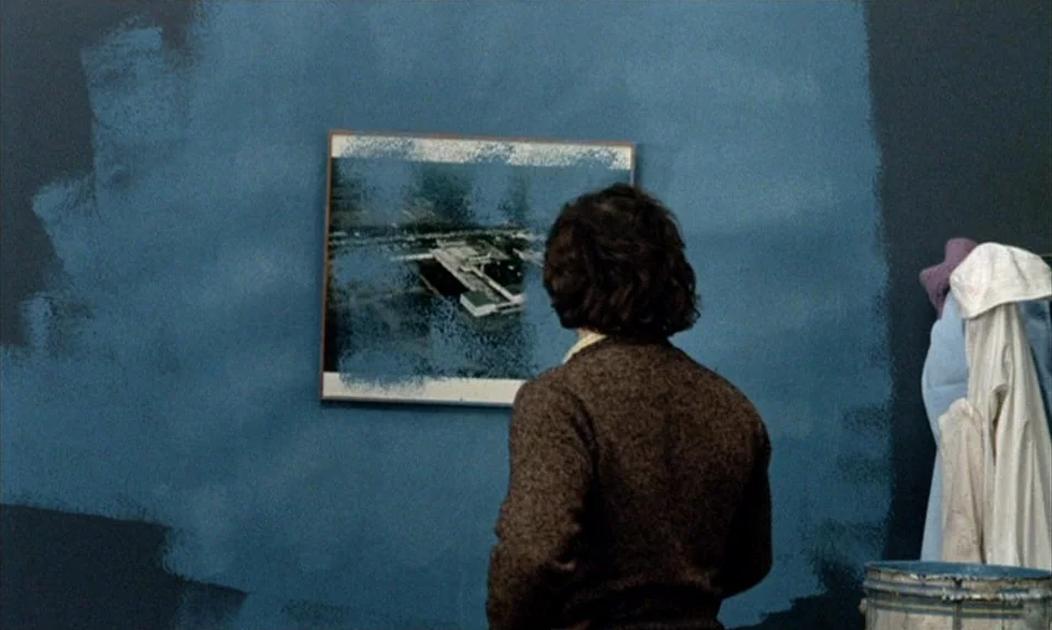 Tout Va Bien (1972) movie still: medium — An interior space features a wall partially painted in uneven streaks of blue paint. A man stands with his…