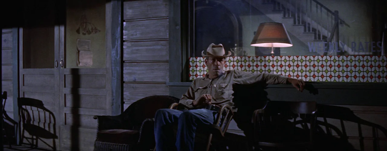 Bad Day At Black Rock (1955) movie still: medium — A wooden porch with a row of chairs sits outside a dimly lit building. A man in a cowboy hat sits…