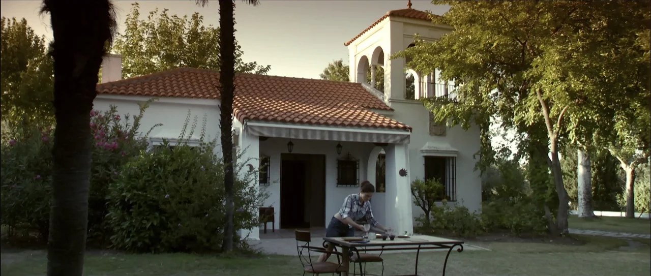 Otro verano (2013) movie still: wide — A white stucco villa with a terracotta roof sits beside a lush green lawn. A woman sets plates on a metal…