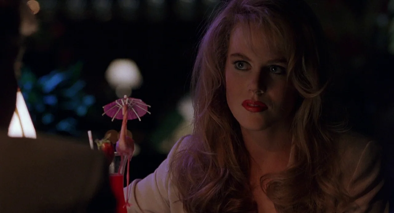 To Die For (1995) movie still: medium — A dimly lit restaurant interior features a woman at a table during nighttime. A red cocktail with a…