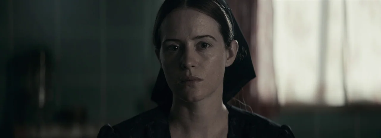 Women Talking (2022) movie still: closeup — A dimly lit room provides the backdrop for a woman in period clothing. She stands still while facing the…