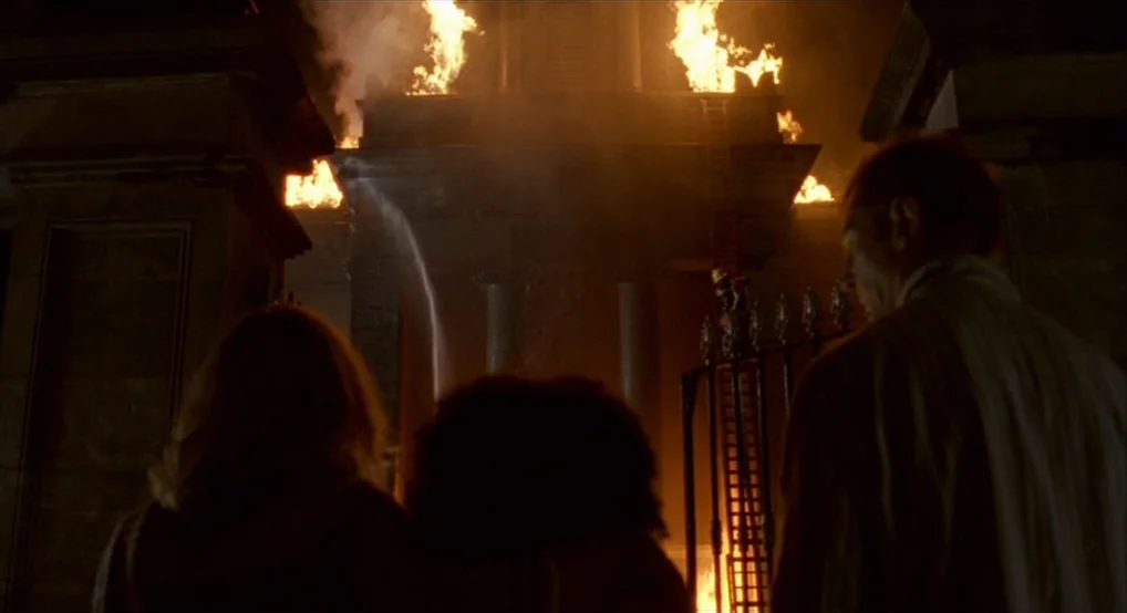 Breakfast On Pluto (2005) movie still: medium — A stone structure burns with bright orange flames against a dark night sky. Three people stand in…
