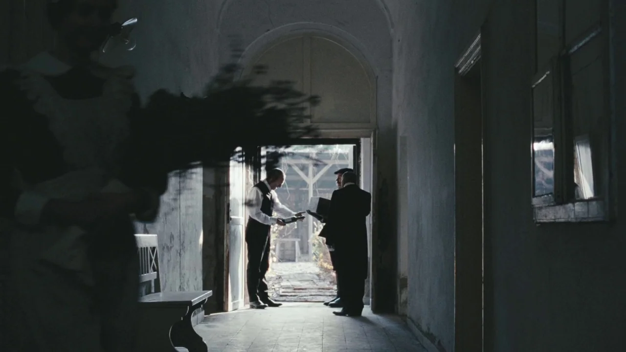 Childhood of a Leader (2015) movie still: wide — A dimly lit stone corridor features an arched doorway leading to an outdoor area. Three men in…