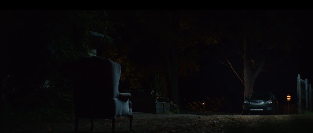 The Front Room (2024) movie still: wide — A dark outdoor space at night features a lone armchair in the foreground. A parked car is visible in the…