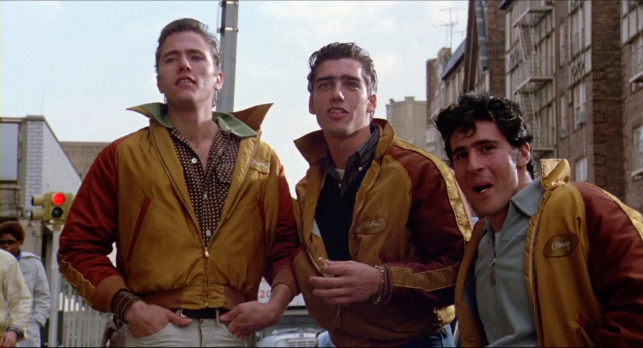 The Wanderers (1979) movie still: medium — Three young men stand together on a sidewalk in front of brick tenement buildings. They wear matching…