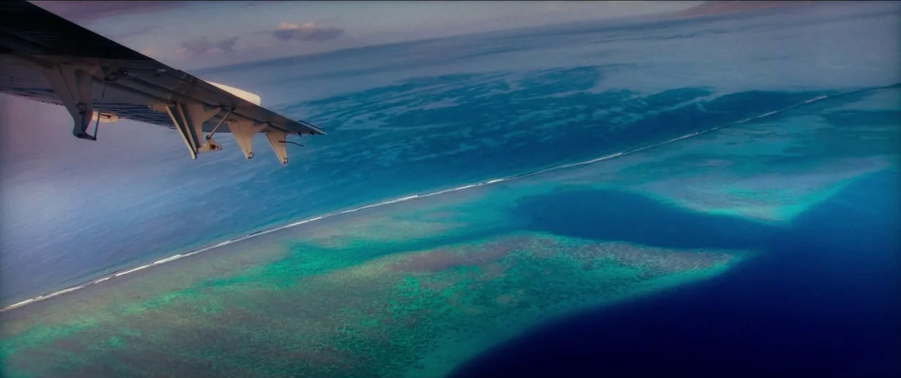 Pacifiction (2022) movie still: wide — An aerial view shows the turquoise waters of a tropical coral reef from above. The wing of a small airplane…