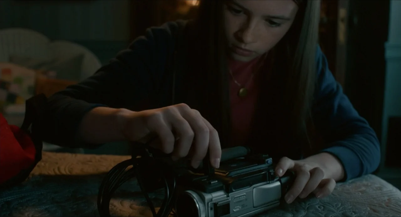 IF (2024) movie still: medium — A dimly lit room features a textured surface where a person works. A teenage girl with brown hair leans over a…