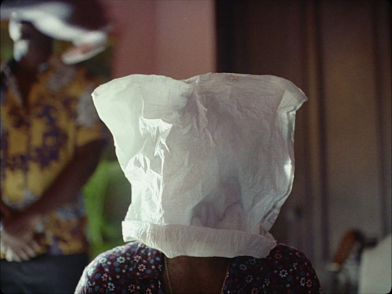 Guava Island (2019) movie still: closeup — A person sits wearing a white woven plastic bag over their head and shoulders. A blurry figure in a…