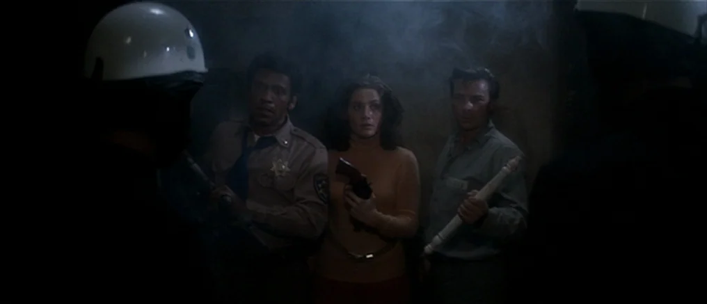Assault on Precinct 13 (1976) movie still: medium — A dimly lit indoor corridor is filled with haze and thick smoke. A police officer, a woman, and…