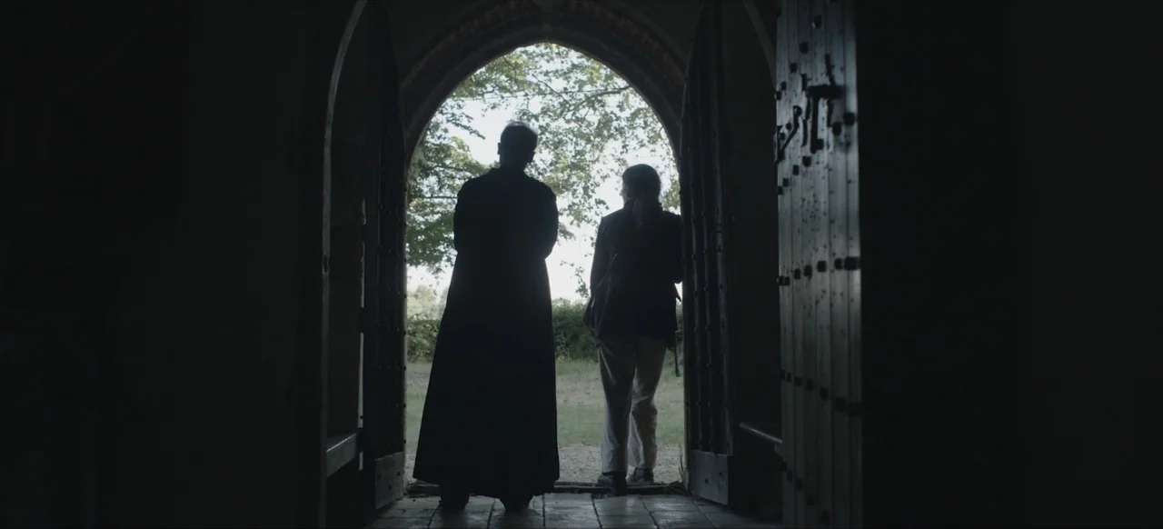 We Go In at Dawn (2020) movie still: fullbody — A stone arched doorway transitions from a dim interior to a bright outdoor area. Two people stand in…