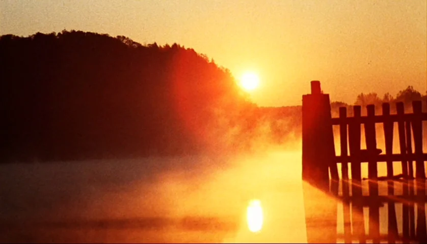 Let’s Scare Jessica To Death (1971) movie still: wide — A wooden pier stands in a calm lake covered in a layer of morning mist. The sun rises behind…
