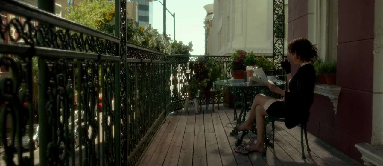 Gerald’s Game (2017) movie still: fullbody — A wrought iron balcony overlooking a city street is filled with potted flowers. A woman sits on a chair…