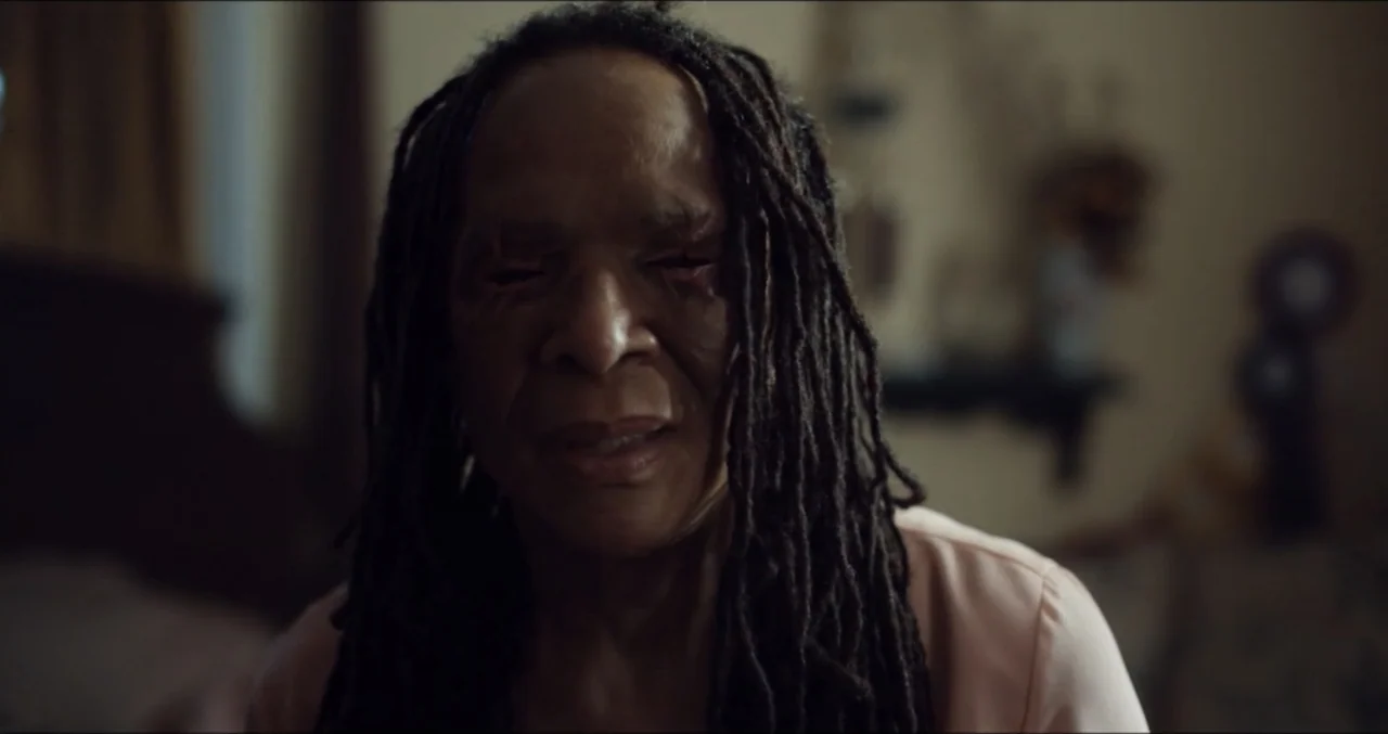 Eye for an Eye (2025) movie still: closeup — A dark, dimly lit bedroom environment with indistinct furniture in the background. An elderly Black…